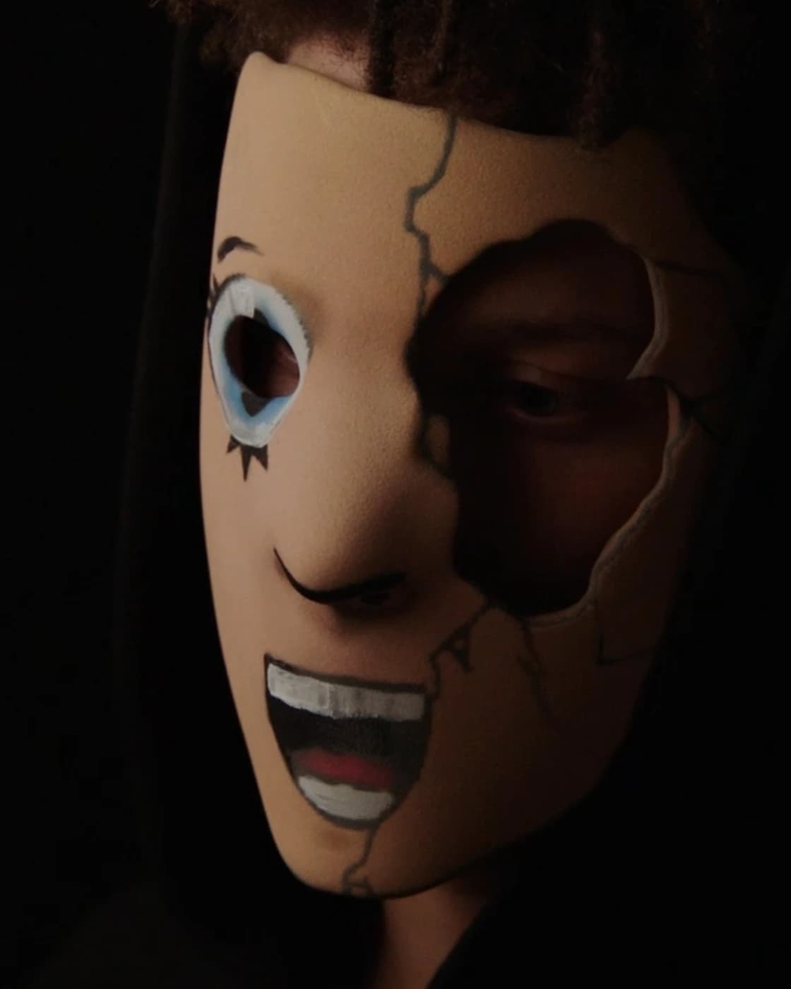 The cracked face worn: one painted eye lit, the hollow cheek in shadow, under a hood
