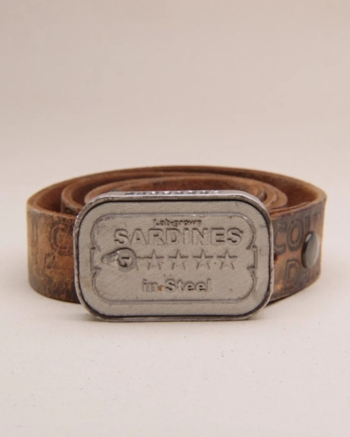 The sardine buckle face-on, the stamped lid reading SARDINES in steel, on a coiled leather belt